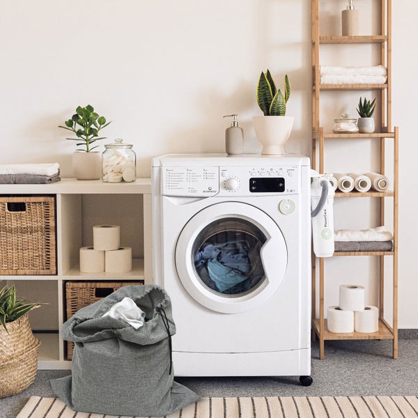 Helle Waschküche mit weißer Waschmaschine, grauem Wäschesack aus Stoff und Holzregalen mit Körben sowie Pflanzen. Organisierte Atmosphäre.