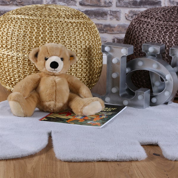 Ein Teddybär sitzt auf einem flauschigen Teppich vor zwei runden Poufs, einem illustrierten Buch und dekorativen beleuchteten Buchstaben.