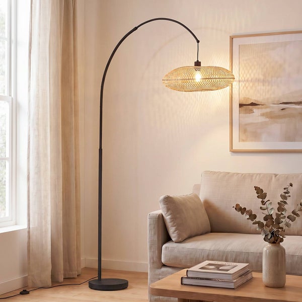Moderne Bogenstehlampe mit Metallgestell und ovalem Schirm aus Geflecht in einem hellen Wohnzimmer mit Sofa und Couchtisch.