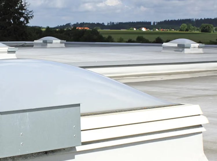 Mehrere kuppelförmige Lichtkuppeln auf einem flachen Industriedach mit Blick auf eine ländliche Umgebung.