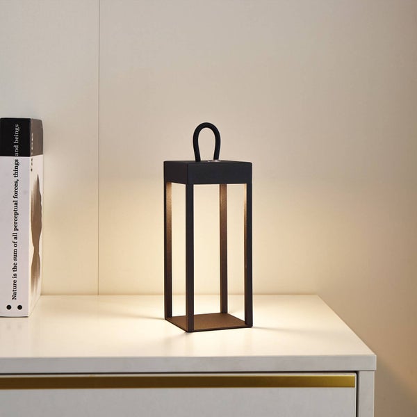 Schwarze LED Tischleuchte in Laternenform aus Metall auf einer weissen Kommode mit warmweissem Licht.