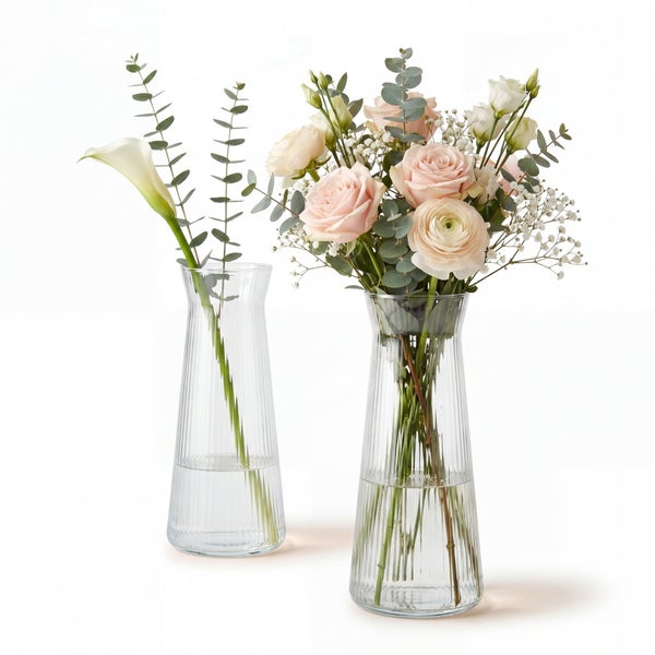 Zwei gerillte Glasvasen, eine mit einer einzelnen Calla und Eukalyptus, die andere mit einem Blumenstrauß aus Rosen, Ranunkeln und Eukalyptus.