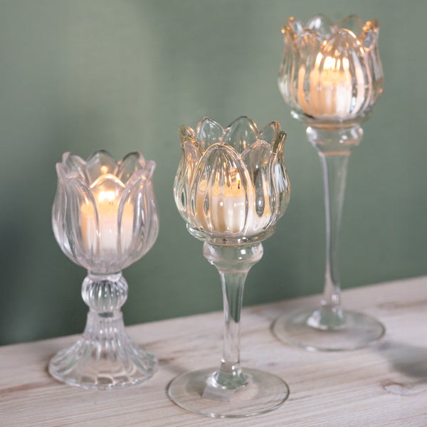 Drei unterschiedlich hohe Teelichthalter aus Glas in Tulpenform mit brennenden Teelichtern auf einem Holztisch.