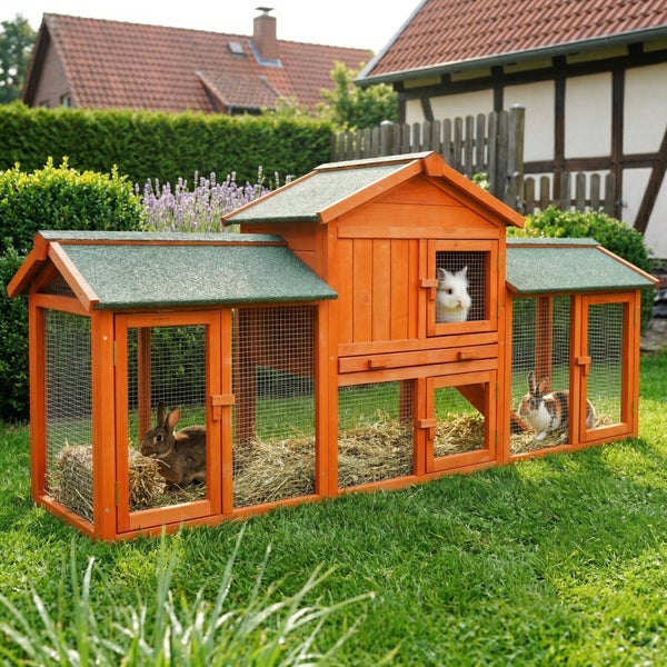 Großer Kleintierstall aus orangefarbenem Holz mit grünem Bitumendach im Garten, drei Kaninchen im Freilaufgehege mit Stroh.
