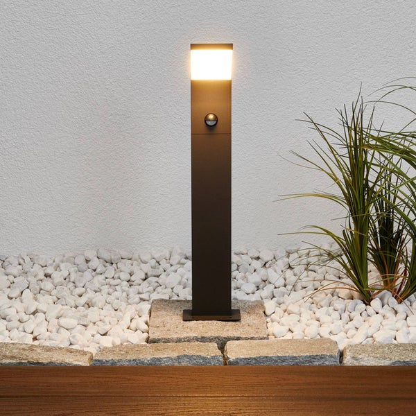 Moderne LED-Wegeleuchte mit Bewegungsmelder, beleuchtet auf Steinfliesen in einem Garten mit Kieselsteinen und Ziergras.