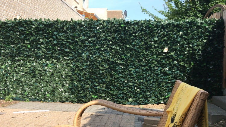 Künstliche Efeuhecke als Sichtschutz auf einer Terrasse im Garten neben einem geflochtenen Korbstuhl.