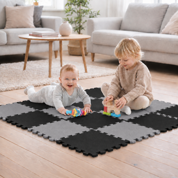 Zwei Kinder spielen im Wohnzimmer auf einer quadratischen Puzzlematte mit einem Schachbrettmuster aus grauen und schwarzen Schaumstoffteilen.