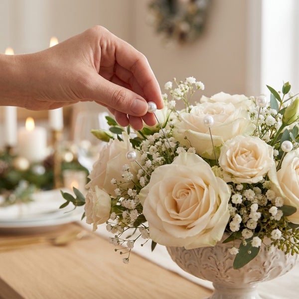 Hand steckt eine Stecknadel mit Perlenkopf in ein Blumenbouquet aus Rosen und Schleierkraut in einer verzierten Schale auf einem gedeckten Tisch.