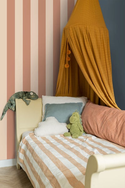 Kinderzimmer mit vertikal gestreifter Tapete, einem Holzbett mit gestreifter Decke, verschiedenen Kissen, Stofftieren und einem Betthimmel aus Stoff.