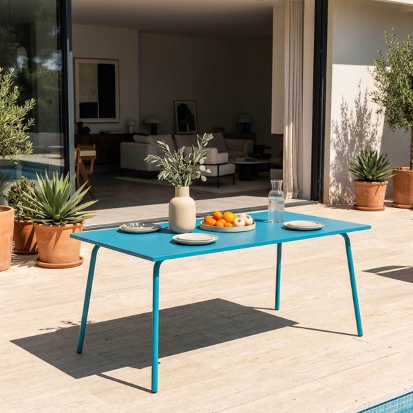 Moderner blauer Gartentisch auf einer hellen Steinterrasse, gedeckt mit Geschirr und Vase, im Hintergrund Pflanzen in Terrakottatöpfen.