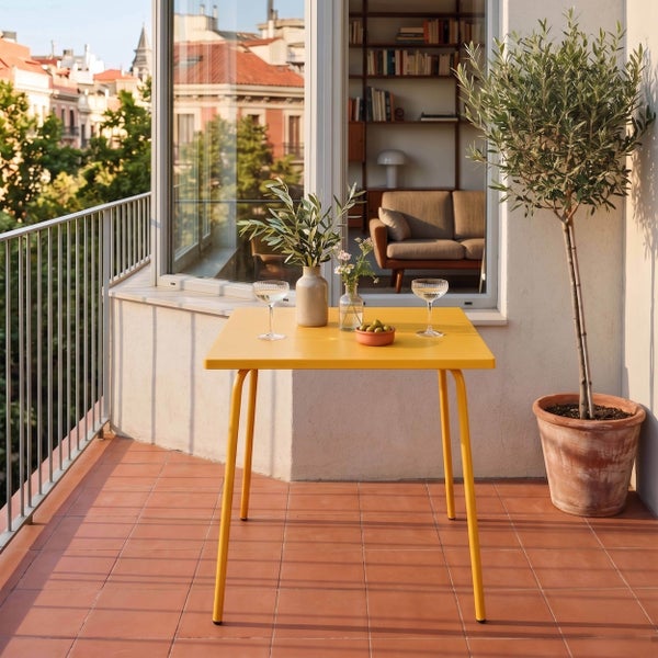 Gelber quadratischer Gartentisch auf einem Balkon mit Terrakottafliesen, Gläsern, Oliven, Vasen und einem Olivenbaum im Topf.