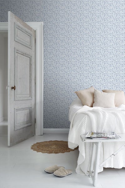 Helles Schlafzimmer mit klein gemusterter Blumentapete, antiker Holztür und Bett mit grob gestrickter Decke.