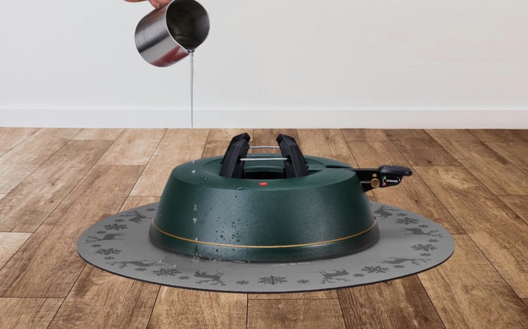 Christbaumständer mit Wasserreservoir auf einer grauen Schutzmatte mit weihnachtlichem Muster, Wasser wird aus einer Kanne eingefüllt.