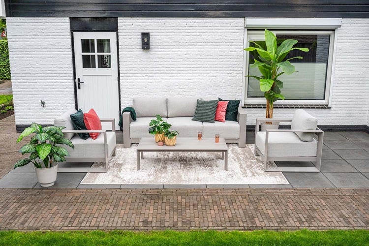 Moderne Gartenlounge aus grauem Aluminium mit Dreisitzer-Sofa, zwei Sesseln und Couchtisch auf einem hellen Teppich vor einer weißen Hauswand.