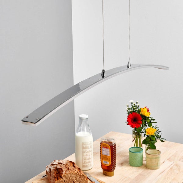 Moderne, gebogene LED-Pendelleuchte in Chrom-Optik über einem Holztisch mit Brot, Milch und Blumen.
