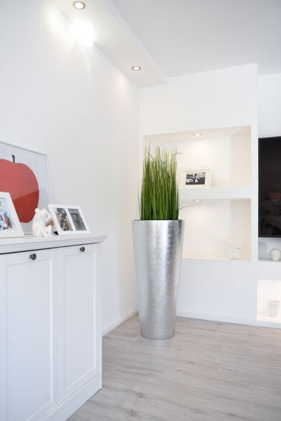 Moderner Wohnraum mit hellem Boden, weißem Sideboard und einer hohen, silbernen Bodenvase mit dekorativem Gras vor einer beleuchteten Wandnische.