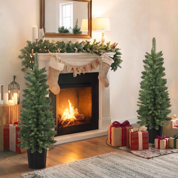 Zwei künstliche Tannenbäume in schwarzen Töpfen vor einem Kamin mit weihnachtlicher Dekoration und Geschenken im Wohnzimmer.