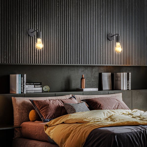 Moderne schwarze Wandablage mit integrierter Leuchte und Vintage-Glühbirne neben einem Bett in einem warm beleuchteten Schlafzimmer.