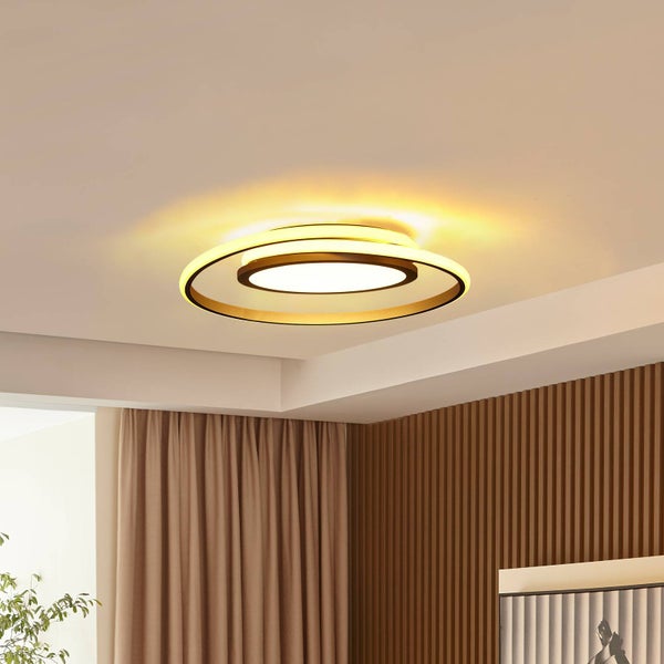 Moderne LED-Deckenleuchte im goldfarbenen Ringdesign in einem hellen Raum mit Holzlamellenwand und Vorhängen.