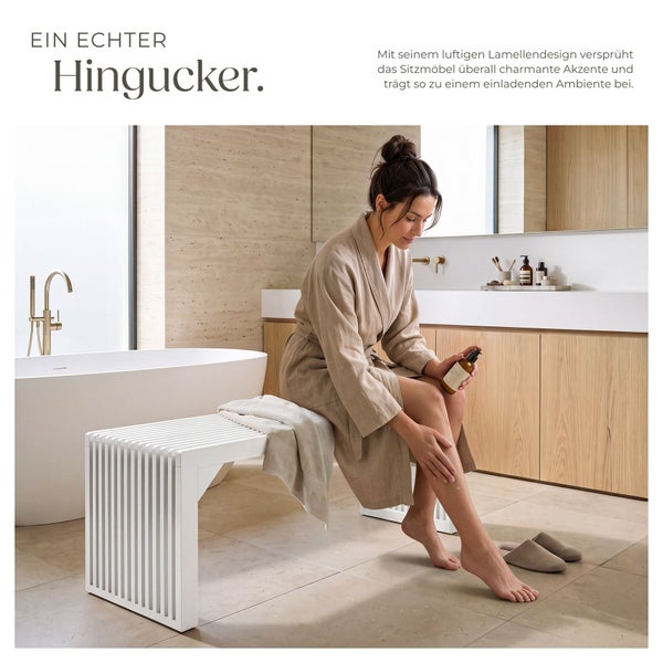 Frau im Bademantel sitzt auf einer weißen Sitzbank im Lamellendesign in einem modernen Badezimmer mit Badewanne und Holzwaschtisch.