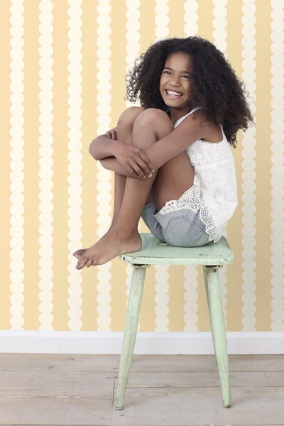 Lächelndes Mädchen sitzt auf einem Hocker vor einer gemusterten Tapete