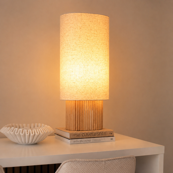 Tischlampe mit zylindrischem Textilschirm und geripptem Holzsockel, stehend auf Büchern neben einer Dekoschale auf einem Tisch.