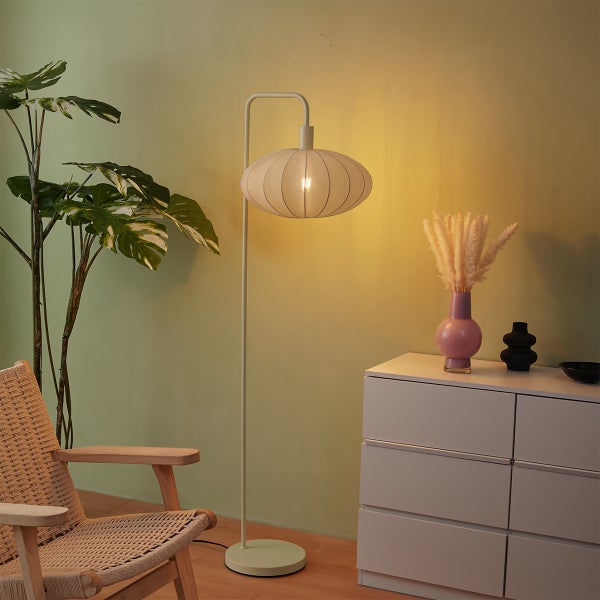 Stehleuchte mit ovalem Stoffschirm und gebogenem Metallgestell in einem hellen Wohnzimmer neben einer Kommode und einem Sessel.