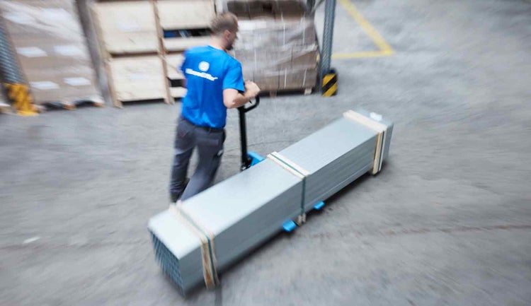 Ein Lagerarbeiter transportiert ein langes Paket mit einem Hubwagen durch ein Logistikzentrum.