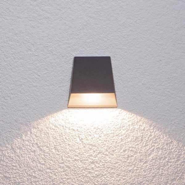 Schwarze Außenwandleuchte in Trapezform mit nach unten gerichtetem Lichtkegel an einer strukturierten Wand.