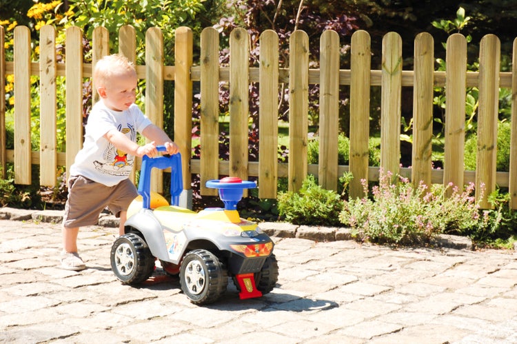 Ein Kind schiebt ein gelb-graues Kinderfahrzeug mit blauen Elementen im Garten vor einem Holzzaun.
