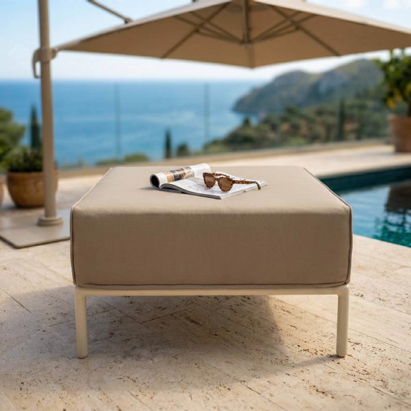 Moderner quadratischer Gartenhocker mit Stoffpolster auf einer Steinterrasse am Pool mit Meerblick unter einem Sonnenschirm.