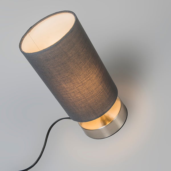 Tischleuchte mit zylindrischem Textilschirm und Metallsockel, eingeschaltet mit warmem Licht.