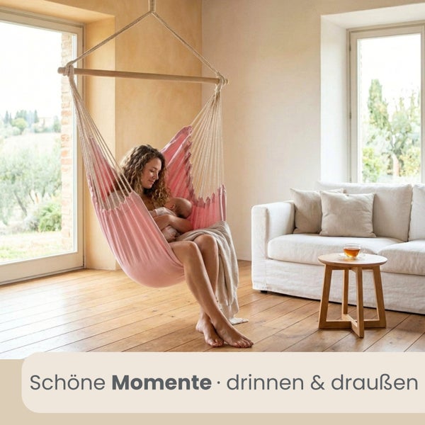 Frau mit Baby in einem Hängesessel aus Stoff mit Holzspreizstab in einem hellen, organisierten Wohnzimmer mit Parkettboden und Sofa.