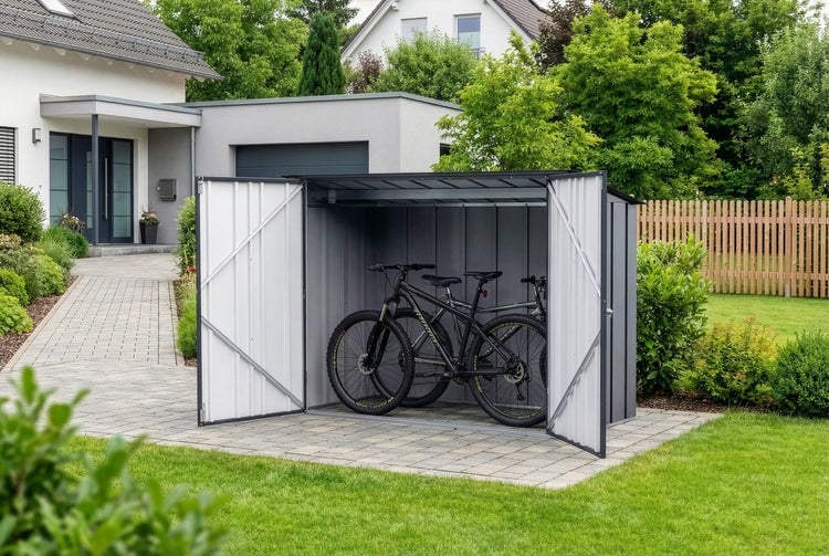 Offene Fahrradbox aus Metall in Anthrazit mit zwei Fahrrädern auf gepflasterter Fläche vor einem Haus im Garten.