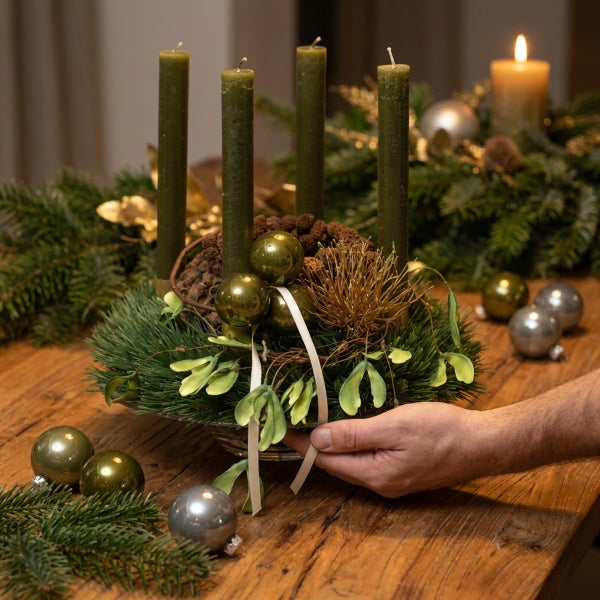 Adventskranz mit vier Stumpenkerzen, Mistelzweigen, Tannengrün und Weihnachtskugeln auf einem Holztisch, gehalten von einer Hand.