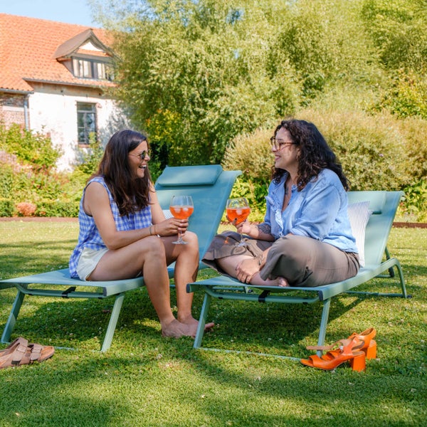 Zwei Frauen entspannen auf hellblauen Gartenliegen aus Metall und Textilgewebe auf einer grünen Wiese im sonnigen Garten.