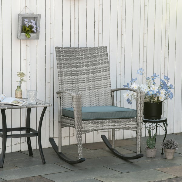 Garten-Schaukelstuhl aus grauem Polyrattan mit Sitzkissen und passendem Beistelltisch vor einem hellen Holzzaun auf einer Steinterrasse.