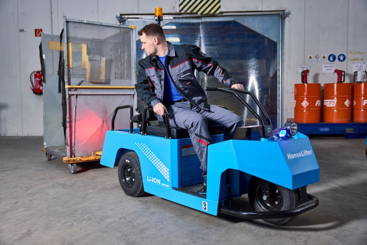 Mitarbeiter bedient einen HanseLifter Elektro-Schlepper mit Lithium-Ionen-Batterie in einem Lager beim Ziehen eines Gitterwagens aus Metall.