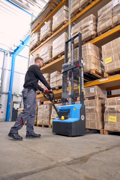 Person bedient einen HanseLifter Elektro-Hochhubwagen in einem Lager mit Hochregalen und Paletten.