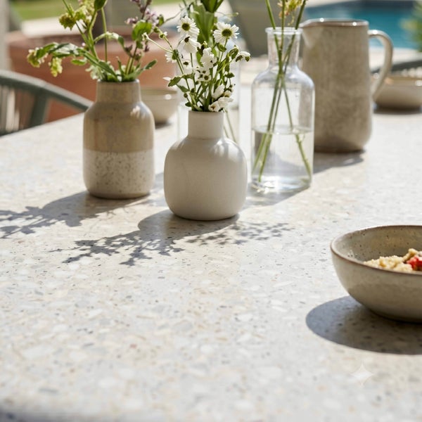 Gartentisch mit Terrazzo-Oberfläche, dekoriert mit kleinen Keramikvasen mit Blumen, einer Glasvase und einem Krug in heller Umgebung.