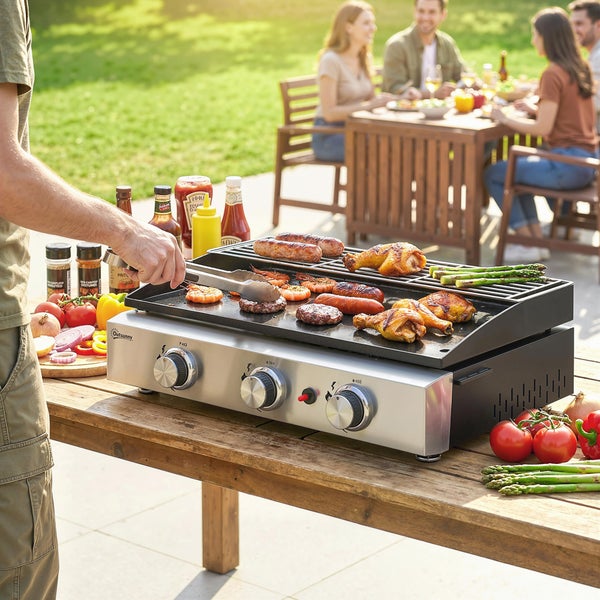 Tischgasgrill aus Edelstahl mit Outsunny Logo auf einem Holztisch im hellen Garten, belegt mit Grillgut, Personen im Hintergrund beim Essen.