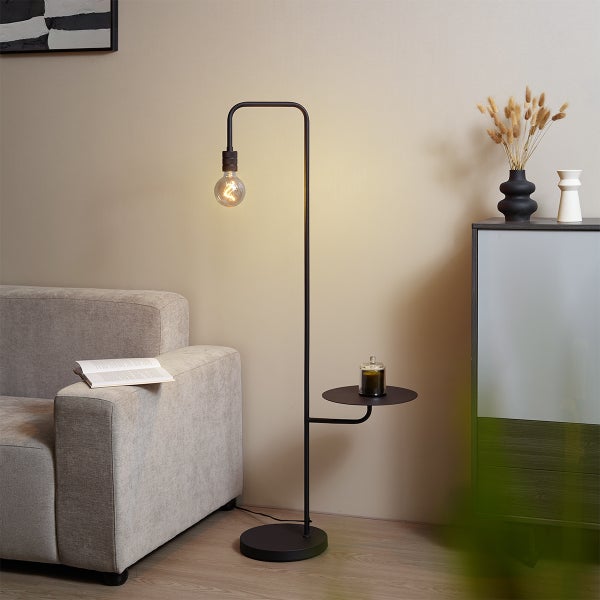 Schwarze Stehleuchte aus Metall mit integrierter Ablage in einem modernen Wohnzimmer neben einem grauen Sofa.