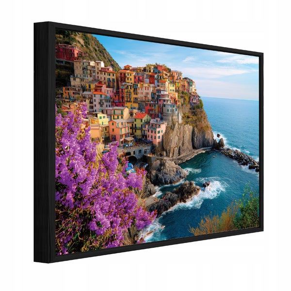Eingerahmtes Wandbild mit Küstenlandschaft, bunten Häusern auf Klippen am Meer und lila Blumen im Vordergrund in schwarzem Rahmen.