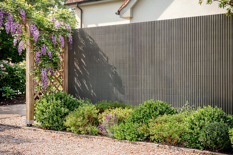Sichtschutzmatte aus Kunststoff zur Montage vor einem Zaun im Garten mit blühendem Blauregen und Sträuchern im Vordergrund