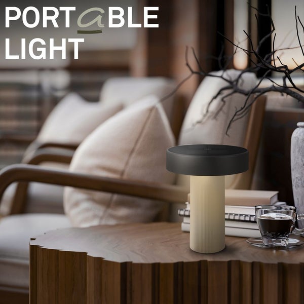 Moderne tragbare Tischleuchte auf einem Holzbeistelltisch in einem gemütlichen Wohnzimmer mit Sesseln und Kissen.