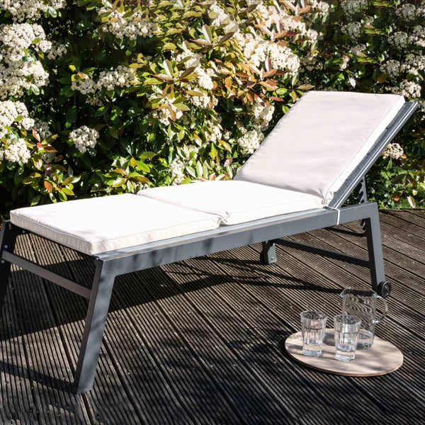Sonnenliege aus Metall mit Polsterauflage auf einer Holzterrasse vor einer blühenden Hecke in einer hellen Atmosphäre.