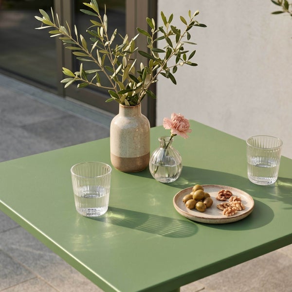 Grüner Gartentisch auf einer Terrasse, dekoriert mit Vasen, Olivenzweigen, Wassergläsern und einem Teller mit Oliven und Nüssen.