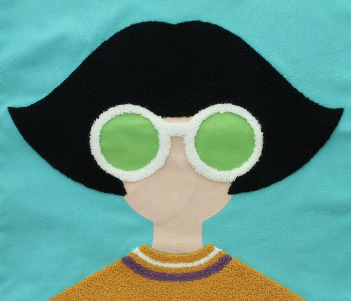 Stoff-Applikation: Charakter mit schwarzem Bob, großer runder Brille und senfgelbem Pullover.