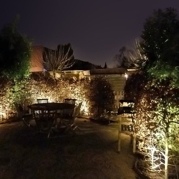 Beleuchteter Garten bei Nacht mit Holztisch, Stühlen sowie durch Bodenstrahler angestrahlten Hecken und Bäumen.