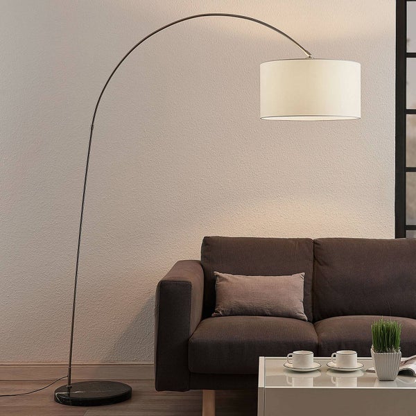 Moderne Bogenstehlampe mit geschwungenem Metallgestell und hellem Stoffschirm hinter einem Sofa in einem hellen Wohnzimmer.